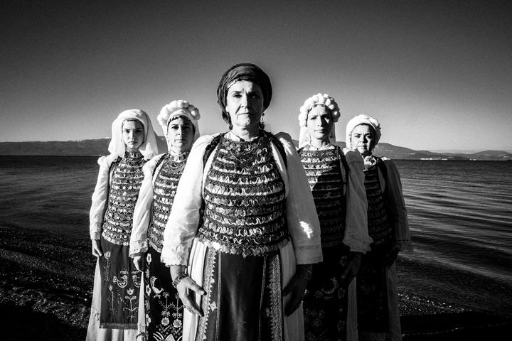 Five women wearing the local costumes of Atalanti, standing in an arrow formation before the sea in Kammena Vourla