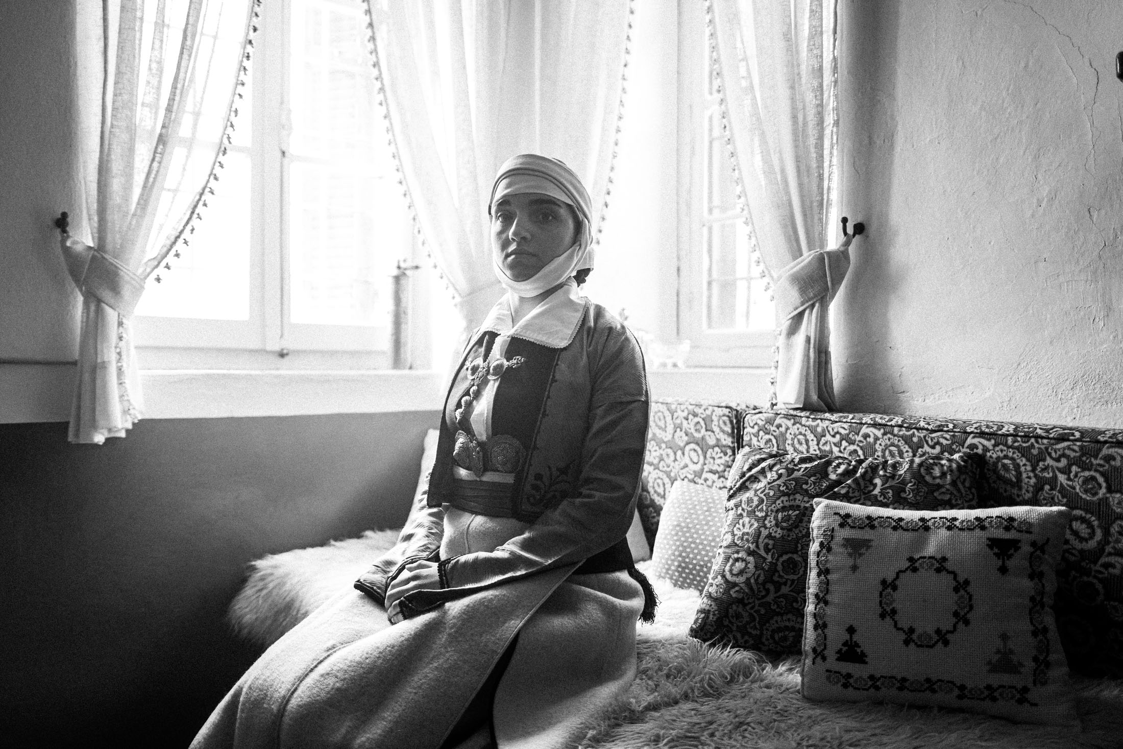 A woman wearing the local costume of Pogoni Epirus, sitting inside an old house in Palaeopyrgos by the window