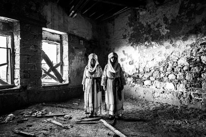 Two women standing side by side, inside an abandoned tannery in the town of Chios, wearing the traditional costumes of Kallamoti