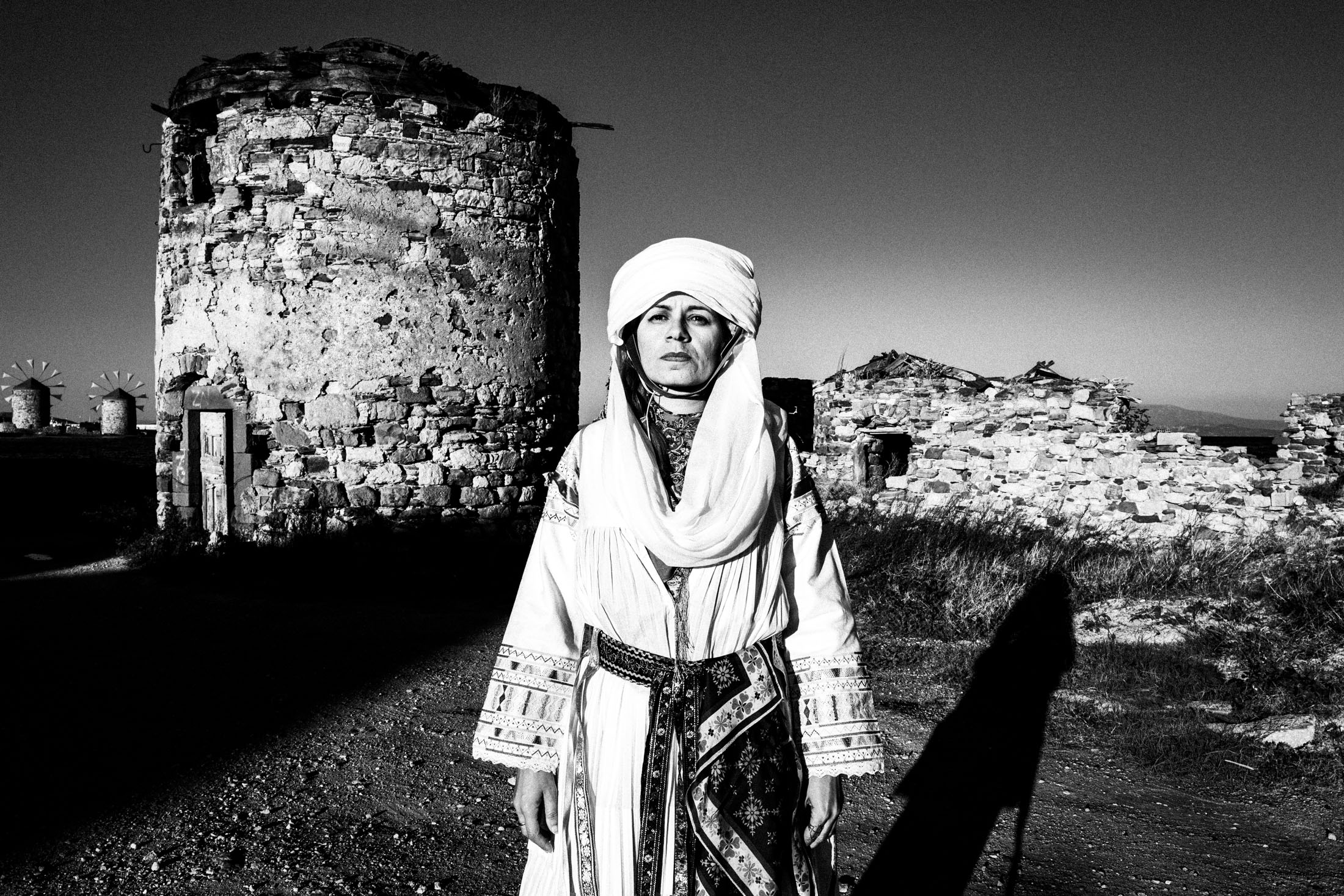 A woman standing in front of the old mills in the town of Chios, wearing the traditional costume of Kallamoti.