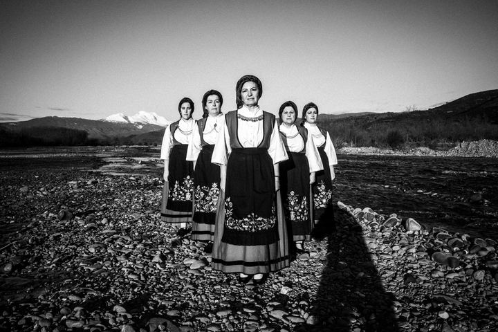 Five women wearing the local costumes of Ypati, standing in an arrow formation by a local river bank, in early morning light