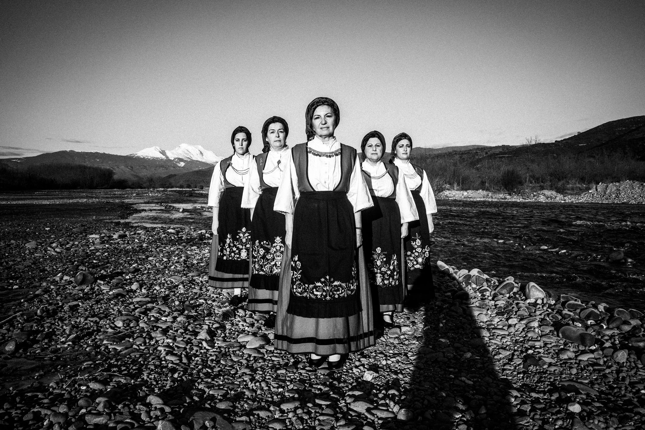Five women wearing the local costumes of Ypati, standing in an arrow formation by a local river bank, in early morning light