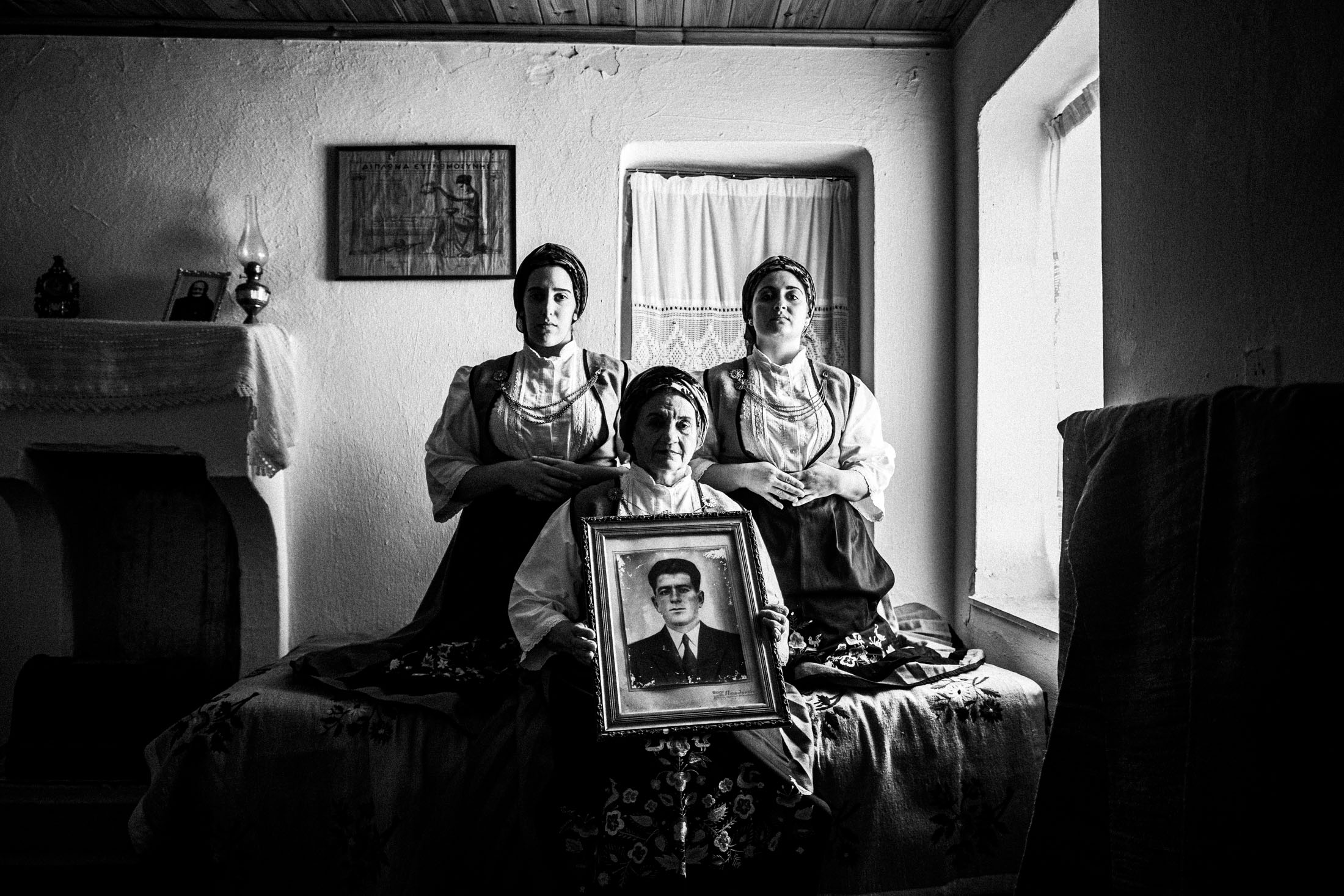 Three women wearing the costumes of Ypati, sitting on a bed holding an old framed photograph at a local house.