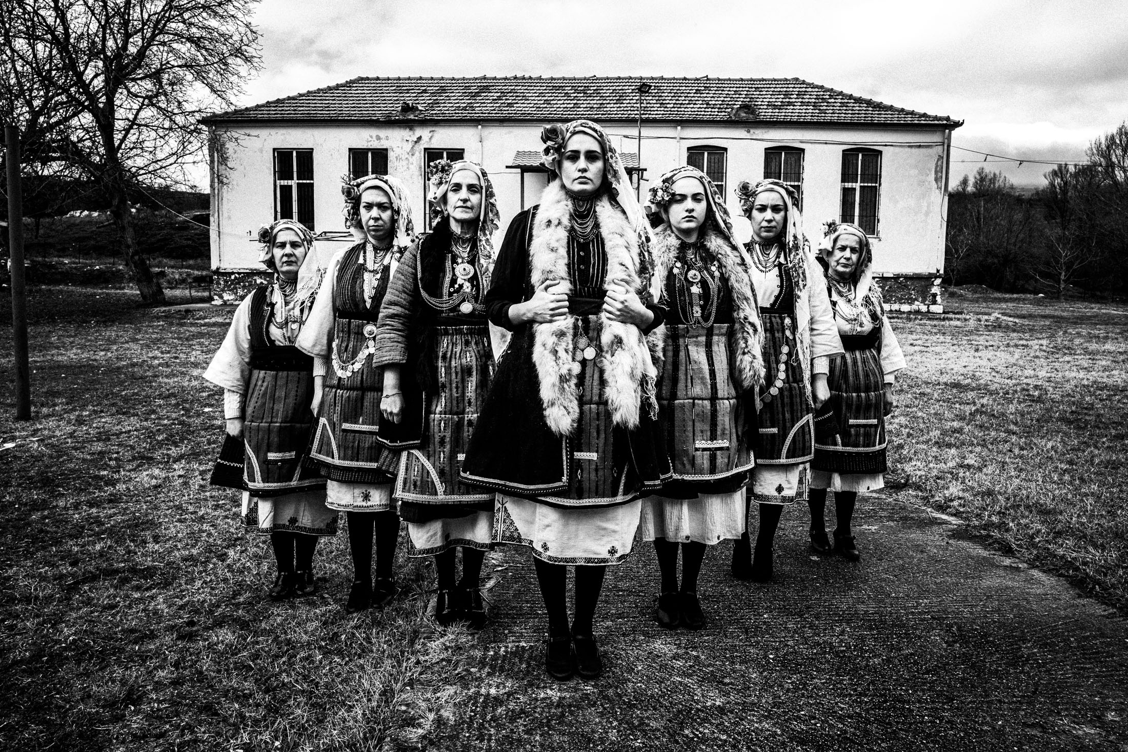 Seven women in the dresses of Skopos, Florina, stand in formation outside the local school that also serves as a folklore museum