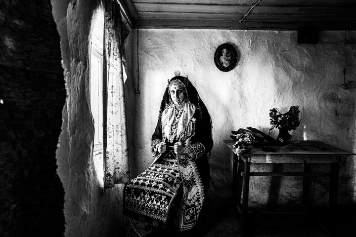 A woman wearing the local dress of Praggi, sitting by the window inside a local home.