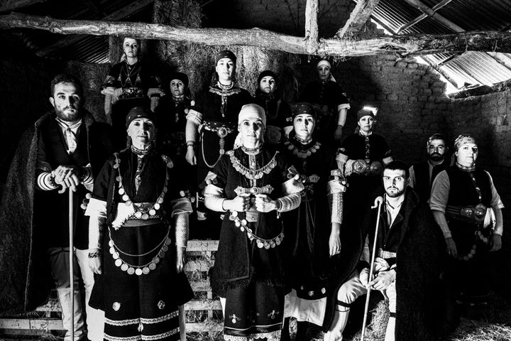 A group of people wearing the local costumes of Prespes lakes, Florina, stand in formation inside a local hay barn
