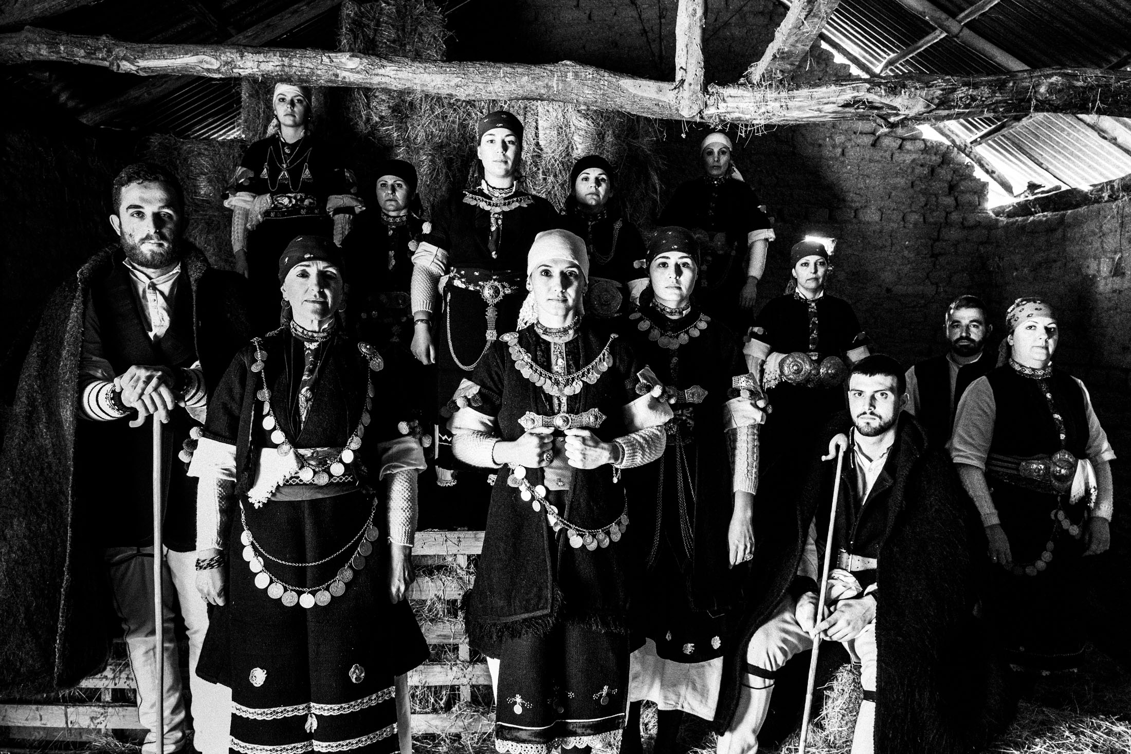 A group of people wearing the local costumes of Prespes lakes, Florina, stand in formation inside a local hay barn