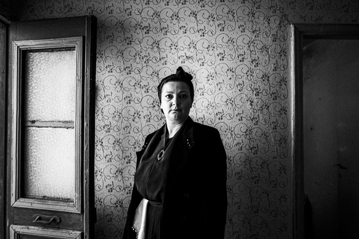 Woman wearing the local costume of Sohos in Macedonia, standing by the door in front of a floral wallpaper, inside a local house