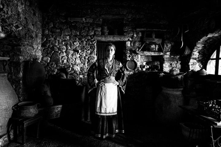 A woman wearing the local dress of Sfakia in Crete, standing inside an old, dark local warehouse.