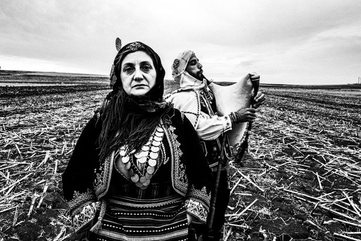 A woman and a man wearing the local costumes of Issaakion in Evros, standing on a ploughed field. The man is playing the traditional bagpipe.