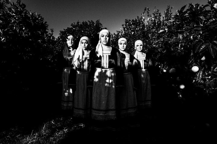 Five women standing in formation amidst a local orange grove, wearing the traditional costumes of Chalkio, Chios.