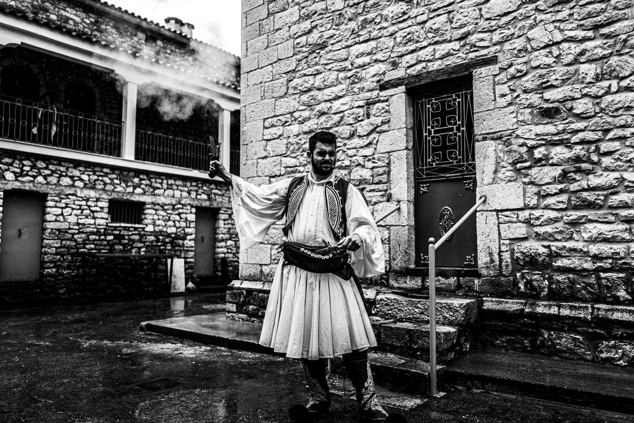 A man in traditional Greek dress firing a historical-style gun loaded with blanks.