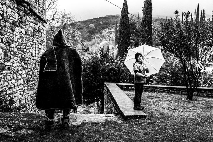 A participant in a heavy traditional coat passing a child with an umbrella at Agia Lavra.