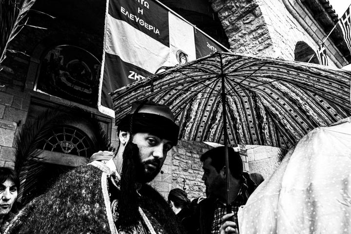A man in traditional dress beneath an umbrella and revolutionary banners at Agia Lavra.