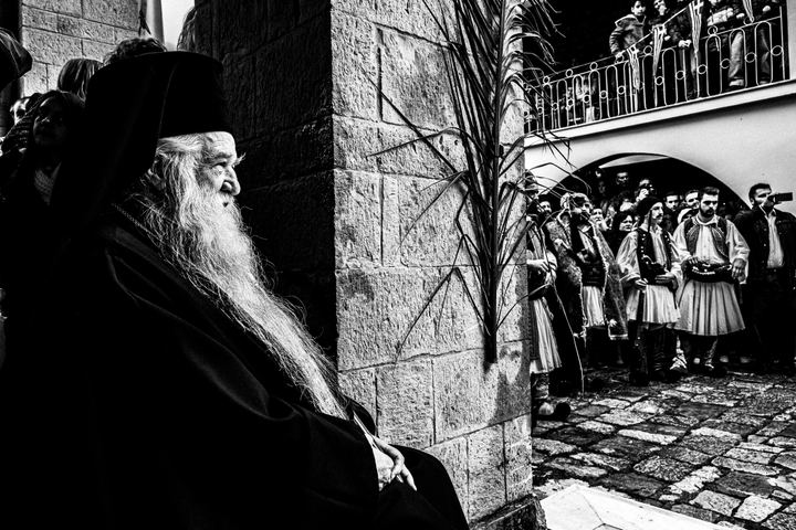 An elderly monk seated beside reenactors in traditional Greek dress at Agia Lavra.
