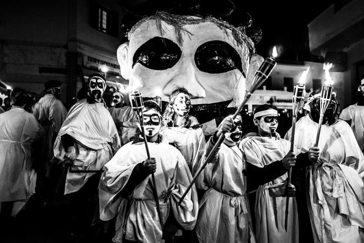 Masked torch bearers gathered beneath the Portara at night.