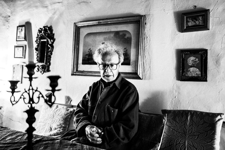 Black and white portrait of Vassilis Kypraios seated indoors, surrounded by framed artworks and antique objects.