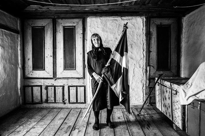 An elderly woman holding a greek flag inside her home at Metaxades
