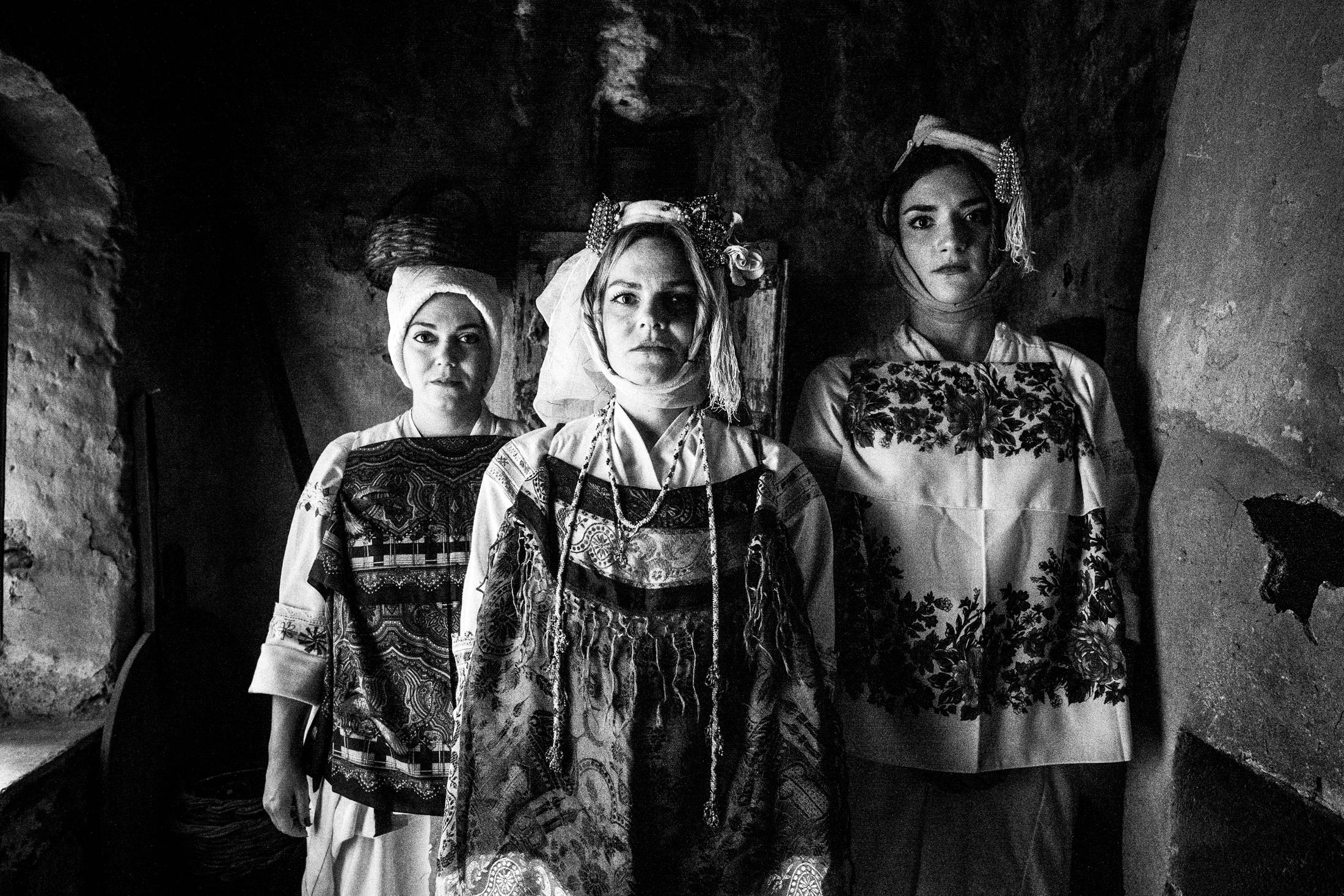 Three women sitting on a bed in an old house, wearing the local dresses of Pyrgi, Chios