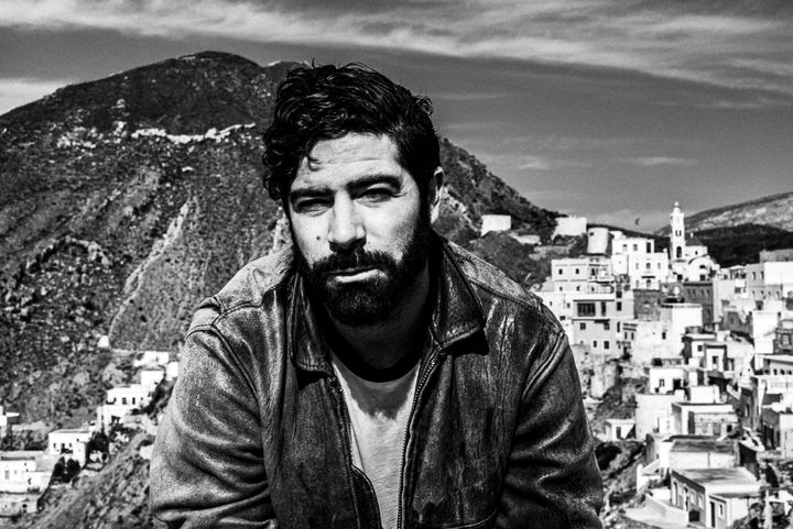 Black and white portrait of Yannis Philippakis with the village of Olympos behind him.