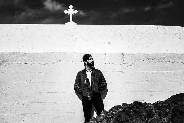 Black and white photograph of Yannis Philippakis standing against a white church wall with a cross above him.