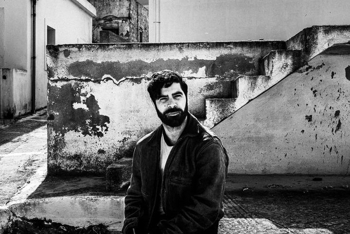 Black and white portrait of Yannis Philippakis seated by whitewashed walls and steps in Olympos.