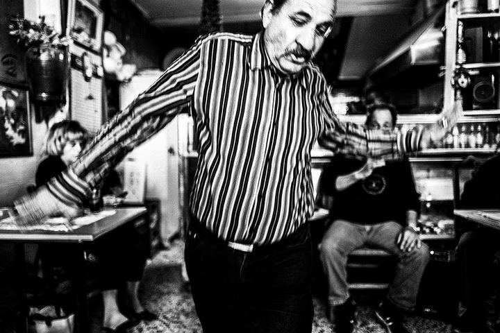 Black and white photograph of a man dancing close to the camera inside Theoklitos Café.