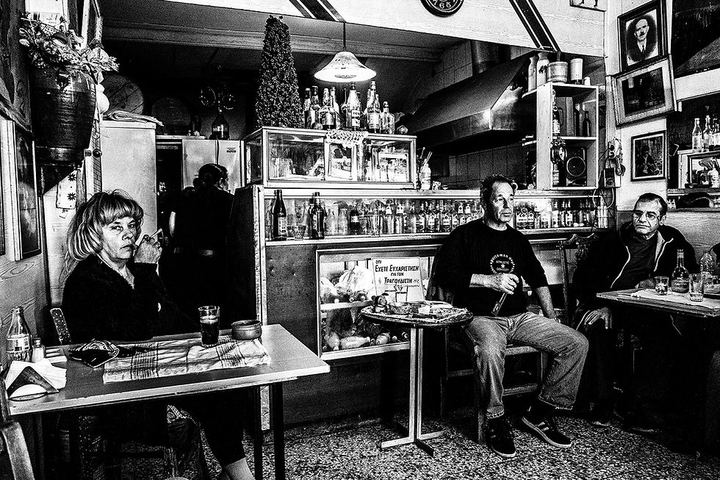 Black and white photograph of the interior of Theoklitos Café, with patrons seated around small tables and Theo holding a microphone.