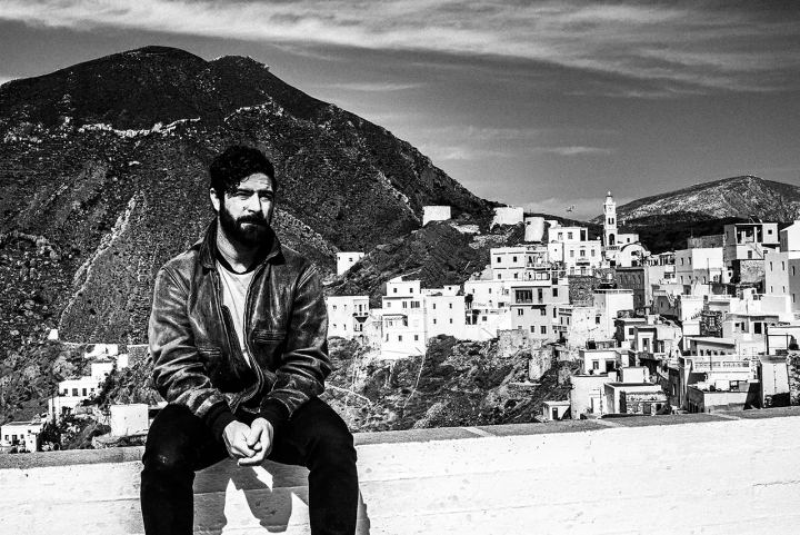 Black and white photograph of Yannis Philippakis seated with the village of Olympos and its church in the background.