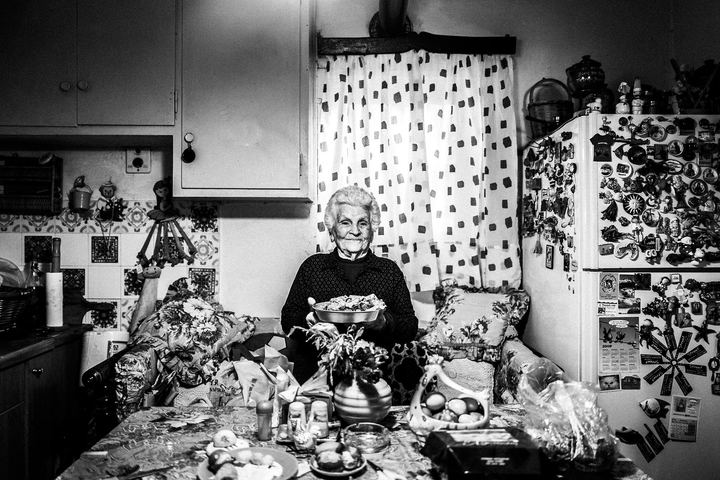Black and white photograph of Chryssi seated at a kitchen table holding a bowl of giaprakia.