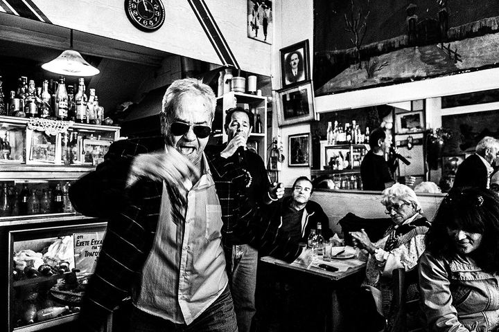 Black and white photograph of a man dancing energetically between the tables of Theoklitos Café.