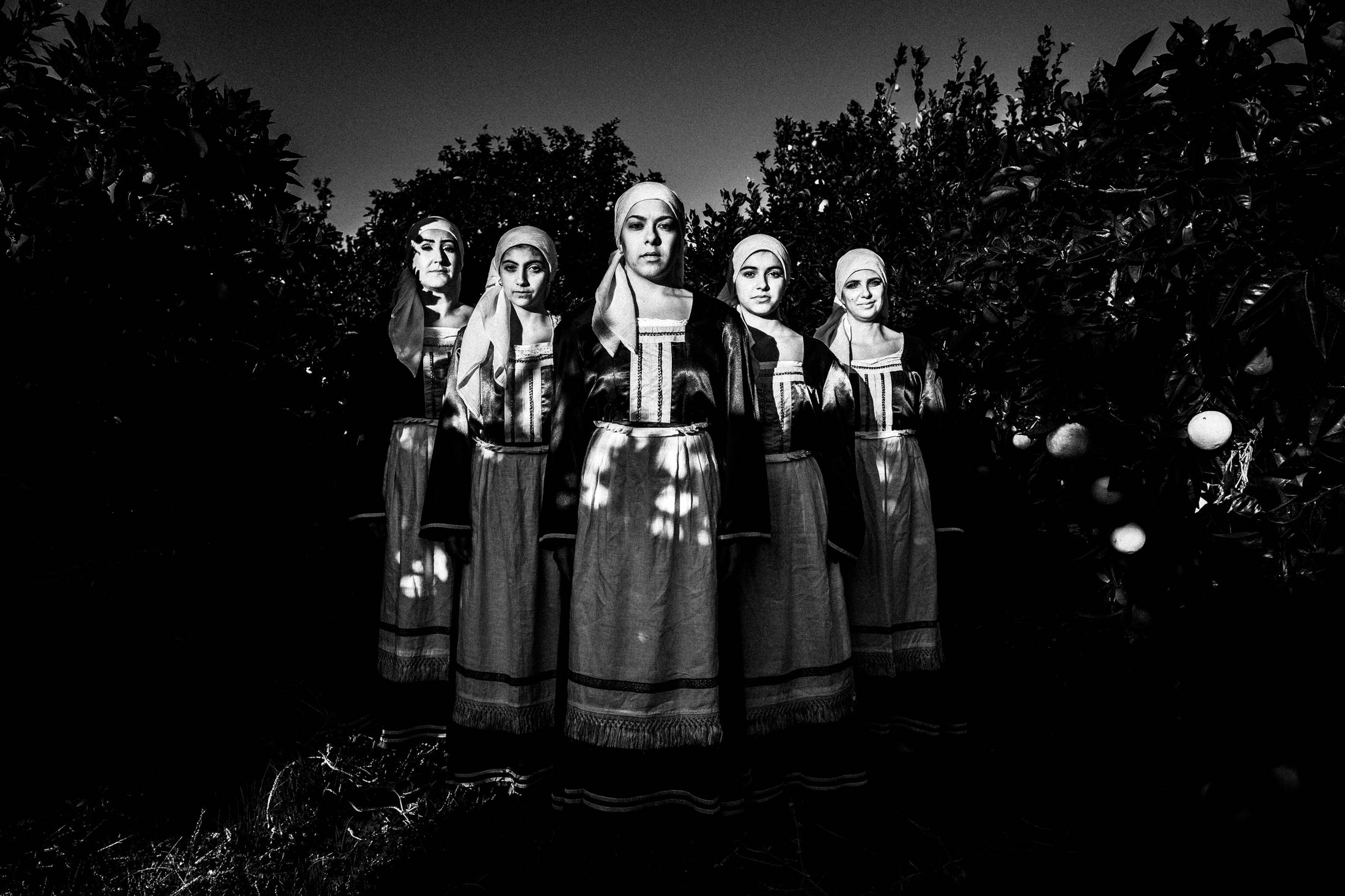 Five women standing in formation amidst a local orange grove, wearing the traditional costumes of Chalkio, Chios.