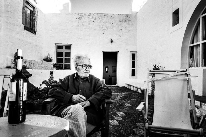 Black and white photograph of Vassilis Kypraios seated in the courtyard of his house in Patmos with a bottle of wine nearby.