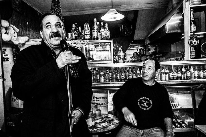 Black and white photograph of a man singing into a microphone while Theo sits beside him in Theoklitos Café.