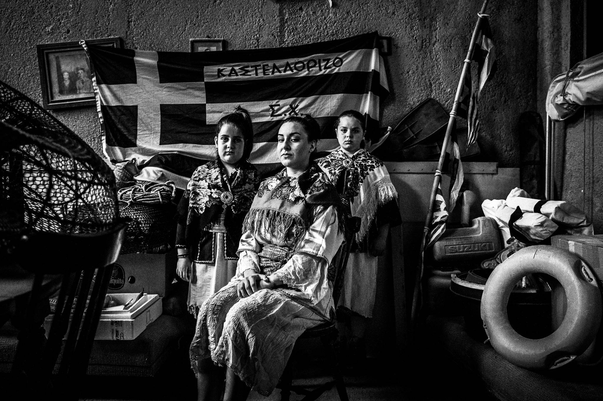 Three young women wearing the costumes of Kastellorizo, posing inside a local fisherman's house with a greek flag behind them
