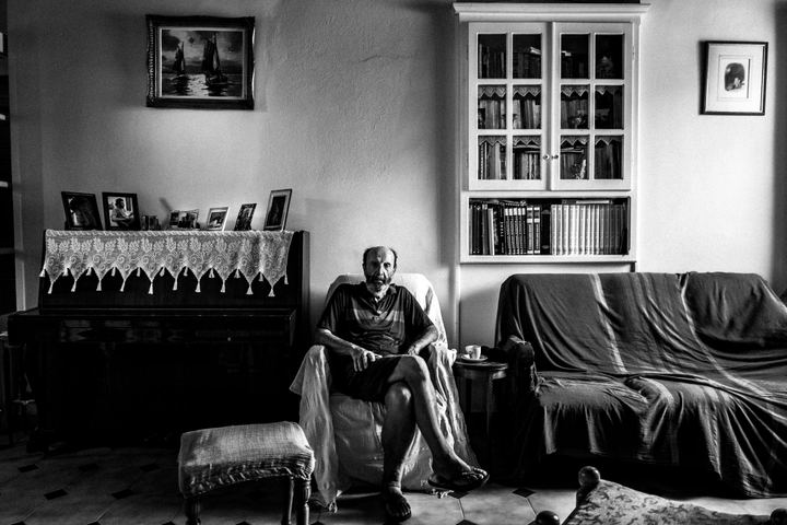 Black-and-white portrait of Dimitrios Kouremenos seated beside a piano in his home on Kalymnos.
