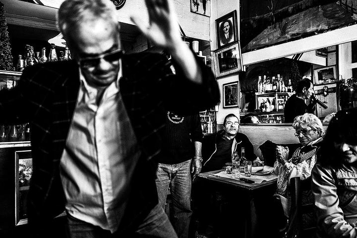 Black and white photograph of a man dancing inside Theoklitos Café while Theo sings in the background.