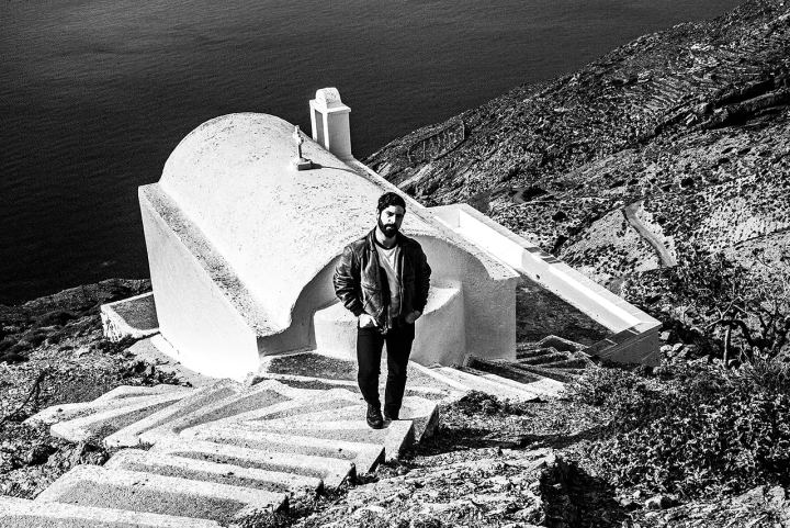 Black and white photograph of Yannis Philippakis standing by a white chapel overlooking the sea near Olympos.