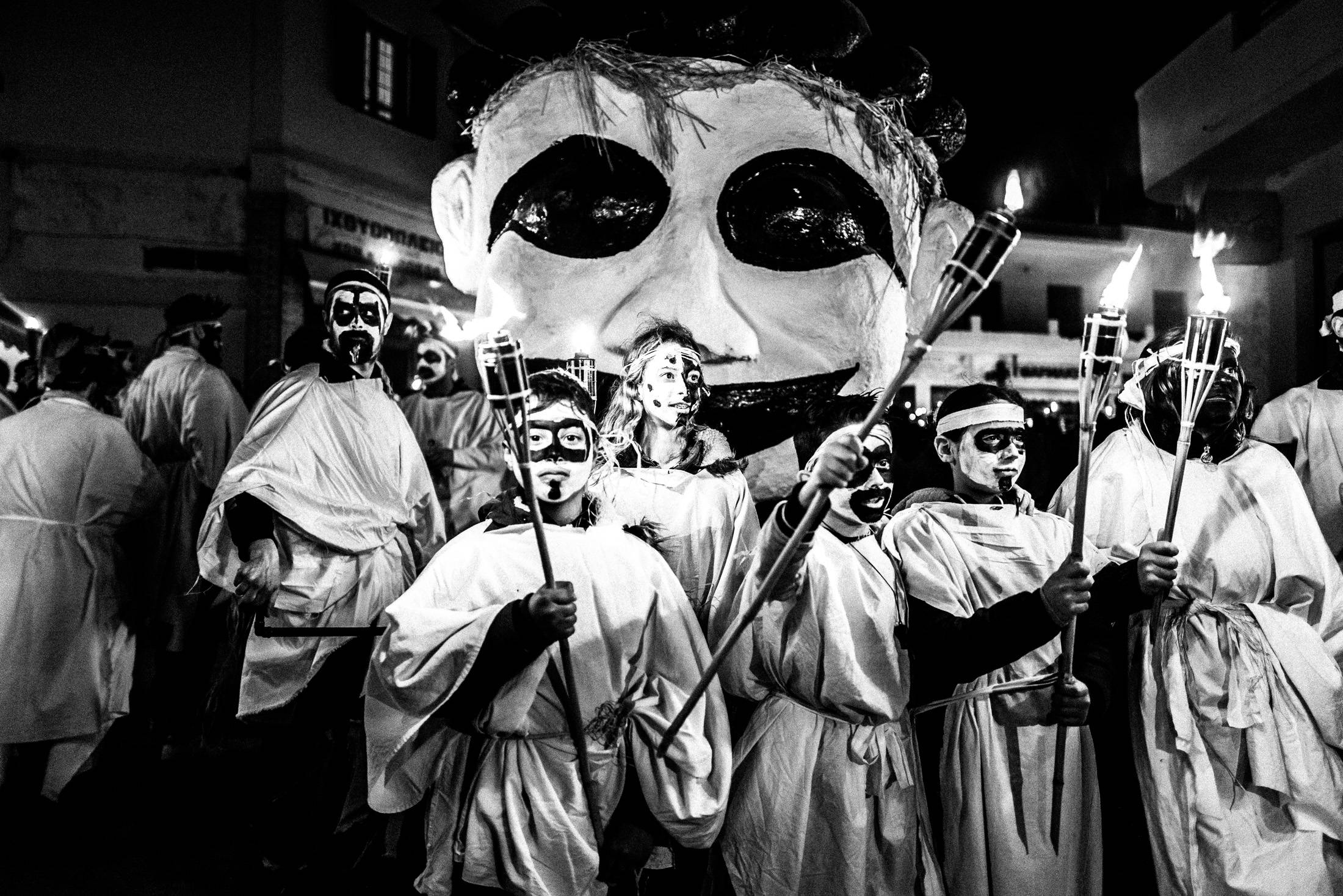 Masked torch bearers gathered beneath the Portara at night.