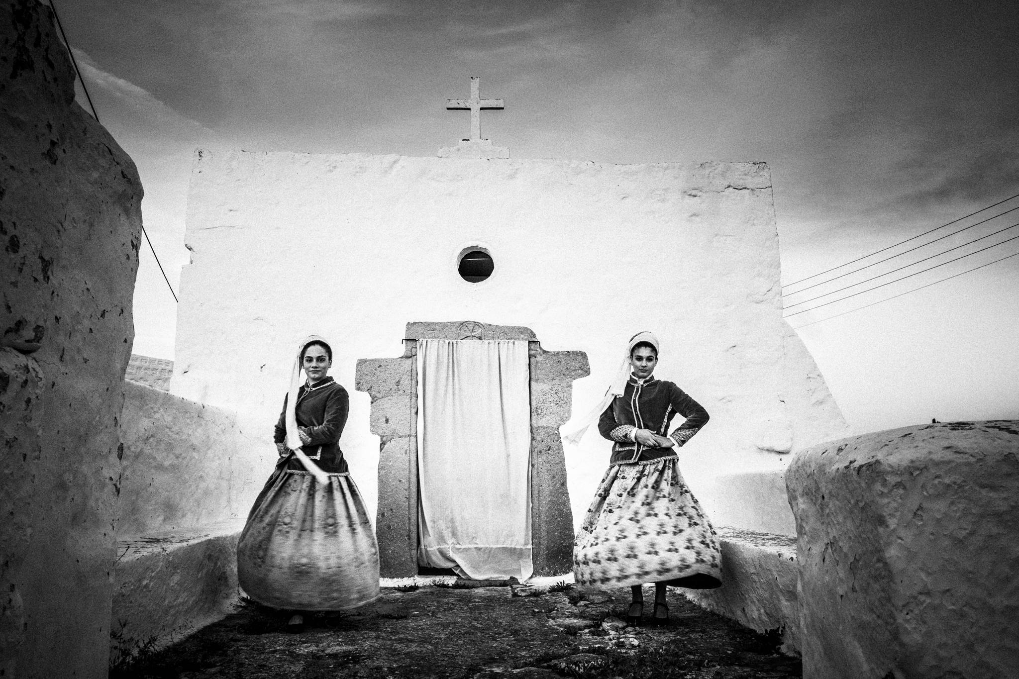Two young women outside a white washed church, wearing the local costumes of Patmos