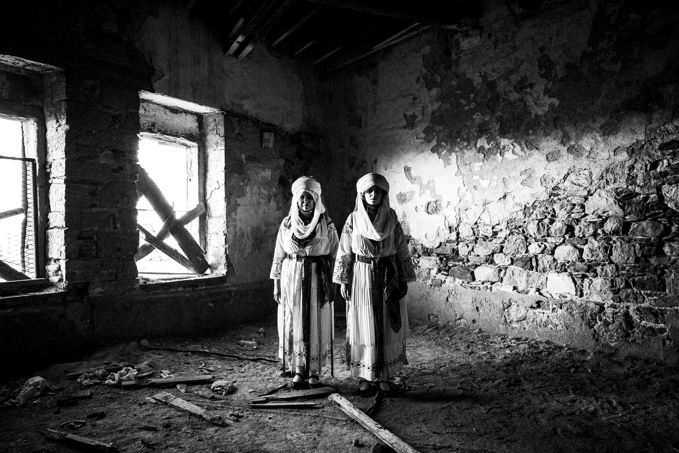 Two women standing side by side, inside an abandoned tannery in the town of Chios, wearing the traditional costumes of Kallamoti