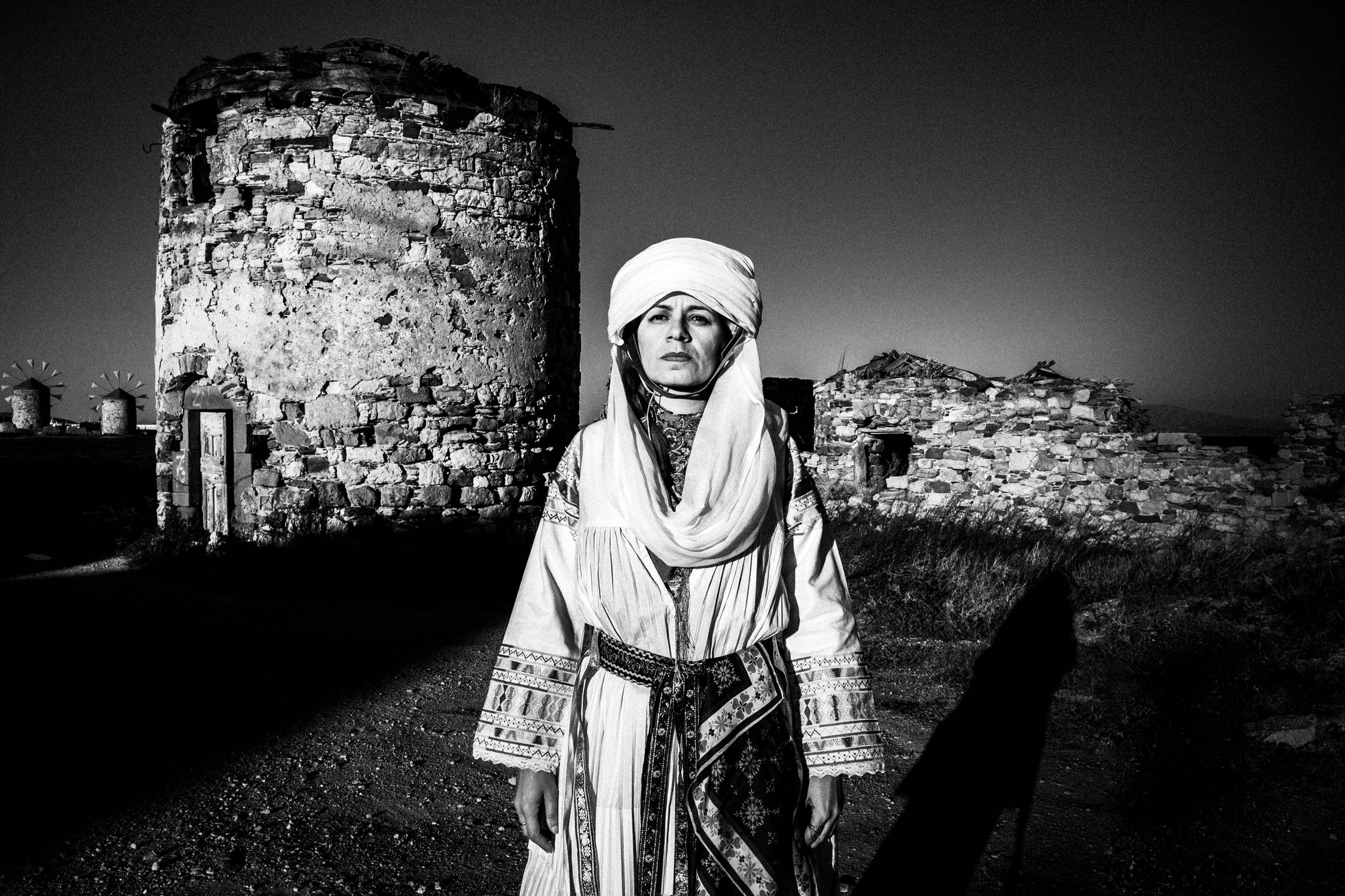 A woman standing in front of the old mills in the town of Chios, wearing the traditional costume of Kallamoti.