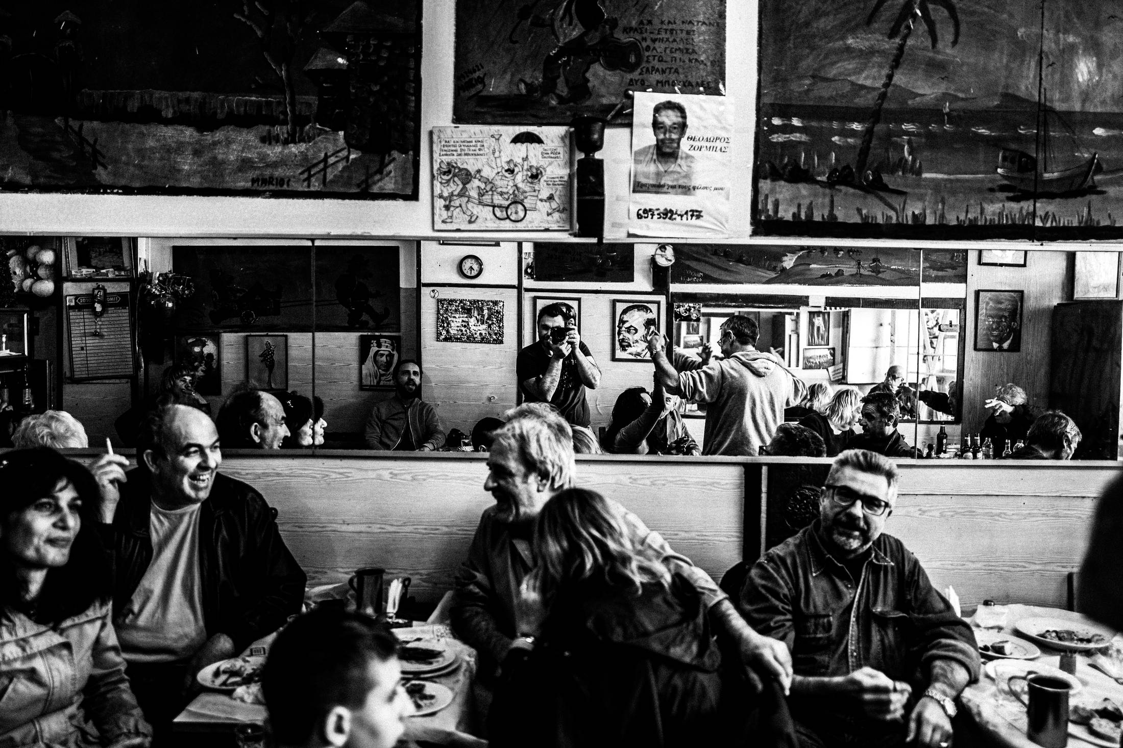 Black and white photograph of people gathered inside Theoklitos Café in Piraeus, with mirrors, paintings and tables full of regulars.