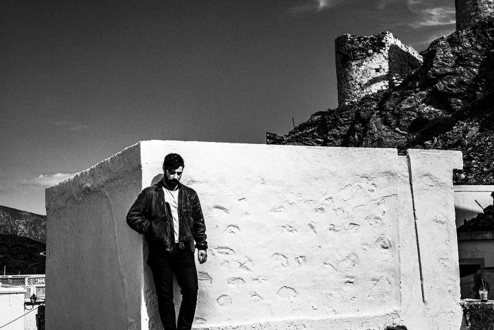 Black and white photograph of Yannis Philippakis standing below stone ruins and whitewashed architecture.
