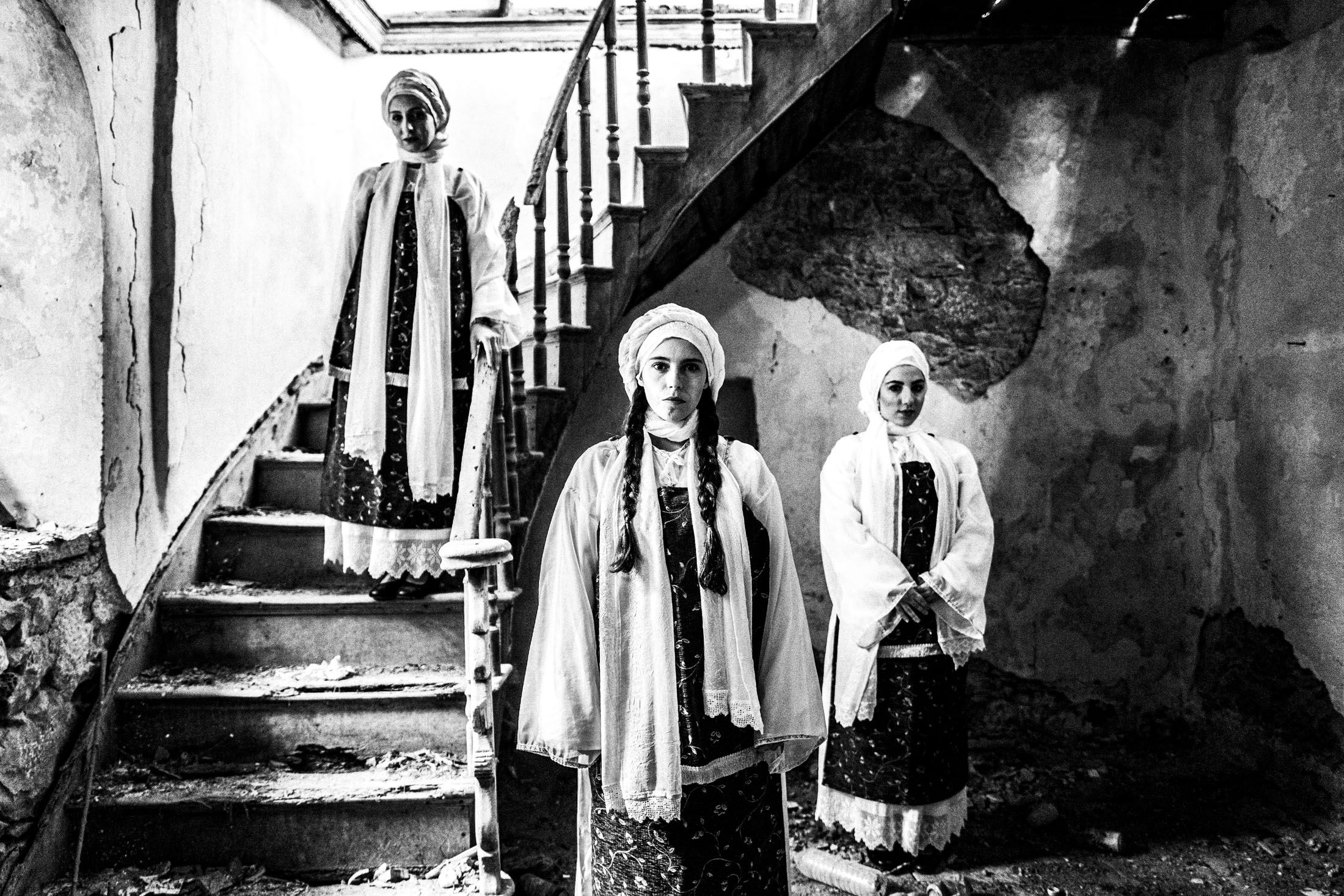 Three women in the dresses of Vessa standing inside an old abandoned house by a staircase